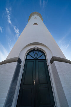 View of Hirtshals fyr lighthouse from a low-angle perspective showing the white tower and dark green entrance door under a blue sky with wispy clouds in Hirtshals, Denmark.