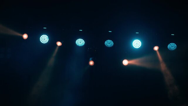 Stage lighting rig with alternating cyan LED par lights and warm orange spotlight heads, thick smoke makes light beams visible against dark teal background