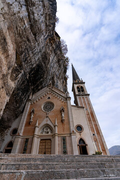 View of Santuario Madonna della Corona built into a vertical rock cliff with a tall bell tower and rose window under a cloudy sky Spiazzi, Veneto, Italy.