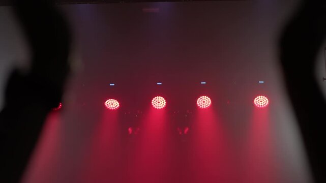 Dark silhouette of raised arms clapping, row of red LED par lights glows behind with smoke diffusing light, dramatic concert atmosphere