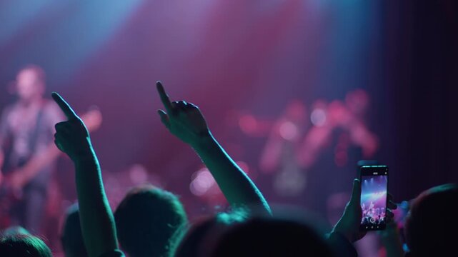 Two hands make rock gesture raised in concert crowd in green light, another fan holds phone to film, red and green stage light visible