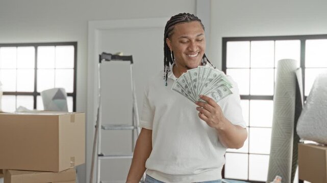 Man holding a fan of dollars, smiling and fanning cash near stacked moving boxes and a ladder inside a building; celebration success.
