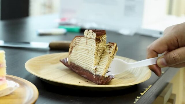 Close up hand using plastic fork cutting slice of chocolate mille crepe cake on wooden plate.