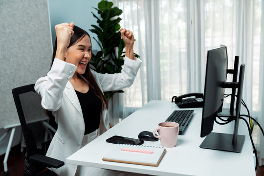 Successful young Asian businesswoman earning highest profit with fist up in exchange stock market on dynamic business graph on pc screens at modern office, analyzing financial technology. Stratagem.