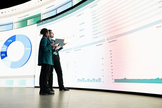 Calculating expenses, graph displayed on videowall and team of investors looking at projected financial statements using laptop. Comparing profit data, charts on display and businessman with colleague