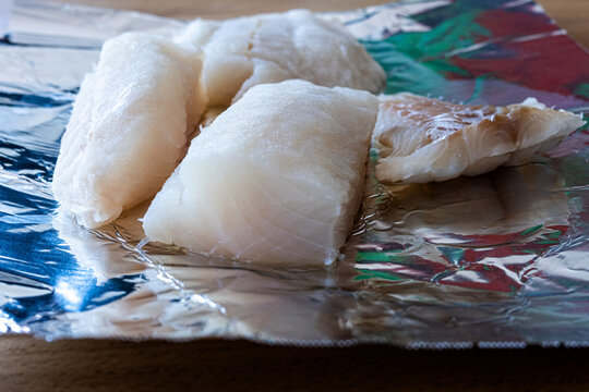 Close-up of multiple pieces of raw, uncooked cod fillets or cod fish loins laid on a tinfoil background. Thick, healthy white fish is being prepared in a restaurant kitchen for frying. Atlantic cod