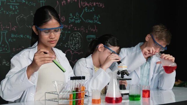 Cute girl looking under microscope while student doing experiment at blackboard with theory written. Young scientist inspect colored solution at table with experimental equipment placed. Edification