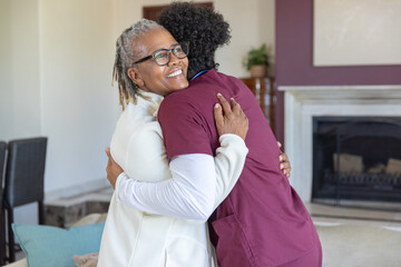 Obraz premium Senior African American woman hugging caregiver wearing maroon scrubs and eyeglasses in living room