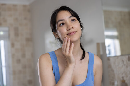 Asian woman wearing blue tank top touching cheek with skincare product at bathroom mirror