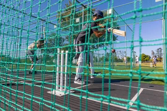 Male cricket players practicing batting in nets with batter wearing helmet pads holding bat stumps