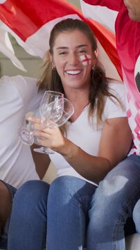 Vertical video: Cheering woman in white tee holding glasses on sofa with England flag after TV goal