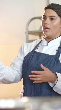 Vertical video: Camera rolling chef coat and cap gesturing explaining prep in kitchen by metal rack