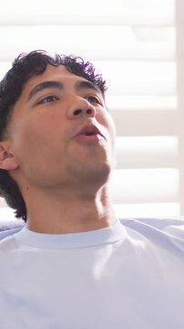 Vertical video: Singing man in blue tee looking up, reclining on home couch by shutters