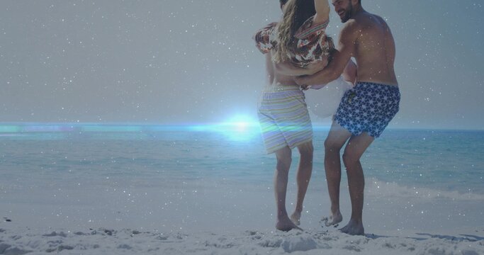 Lifting parents holding child at shoreline with footprints and sun glare, in swim trunks and sarong