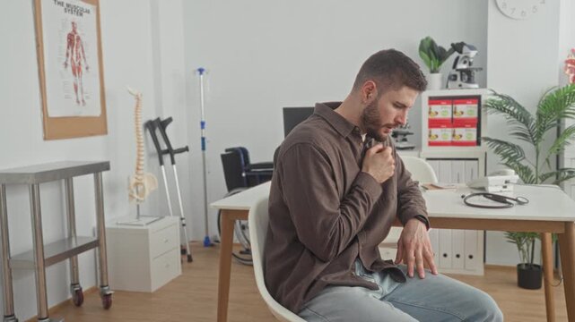 Man clutches chest and coughs into fist while seated at a medical desk with stethoscope and spine model in clinic building, suggesting possible heartattack; anxiety urgency.