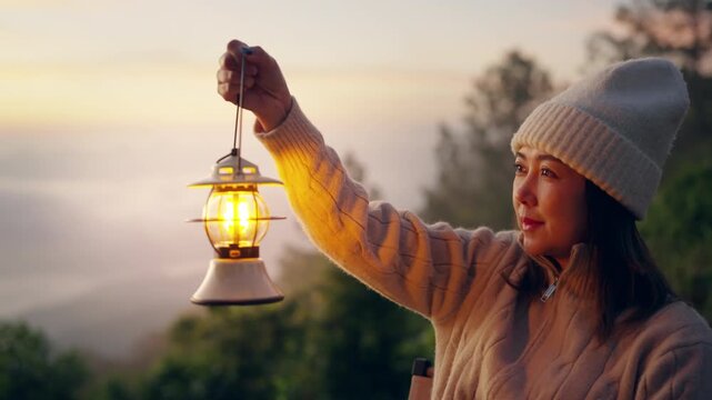 Asian woman holding camping lantern at sunrise mountain peak, female traveler enjoy peaceful morning with nature therapy, outdoor travel active lifestyle, hiking and camping trip with cloudscape sky