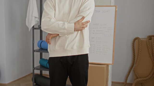 Man holds left elbow in a fitness studio with whiteboard, foam rollers and battle rope visible; calm reflection.
