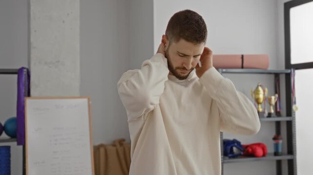 Man rubbing neck and massaging nape for neck pain in a fitness studio with yoga mat and boxing gloves visible; discomfort recovery.