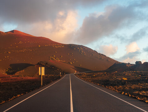 View of an empty asphalt road winding through the volcanic landscape of Timanfaya National Park with reddish mountains under a soft sunset sky Lanzarote, Canarias, Spain.