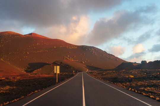 View of a paved road winding through the volcanic landscape of Timanfaya National Park with red hills and lava fields under a soft sunset sky Lanzarote, Canarias, Spain.