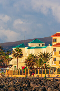 View of a coastal resort with turquoise roofs and palm trees as people walk along the promenade against a mountain backdrop in Lanzarote, Canarias, Spain.
