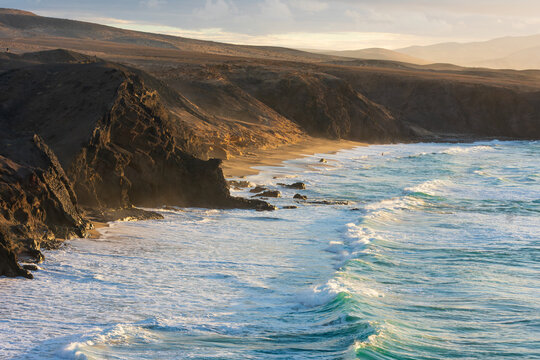 View of volcanic cliffs overlooking a sandy beach with turquoise waves crashing on the shore under warm golden light Lanzarote, Canarias, Spain.
