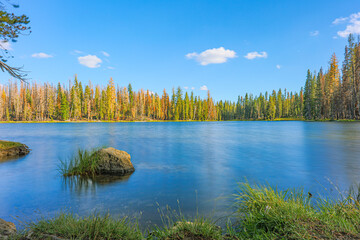 A smooth blue alpine lake with a boulder and grassy shore reflects autumn orange and green conifers under a partly cloudy sky in Lassen Volcanic NP, CA. © ZL Visuals