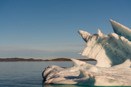 View of a large iceberg with sharp jagged peaks floating in the calm blue waters of a fjord with distant rocky hills under a clear sky in Nuuk, Sermersooq Municipality, Greenland.