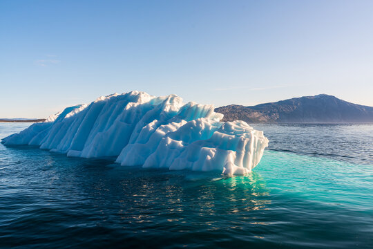 View of a large jagged iceberg with bright blue textures floating in the ocean near rocky mountains in Nuuk, Sermersooq Municipality, Greenland.