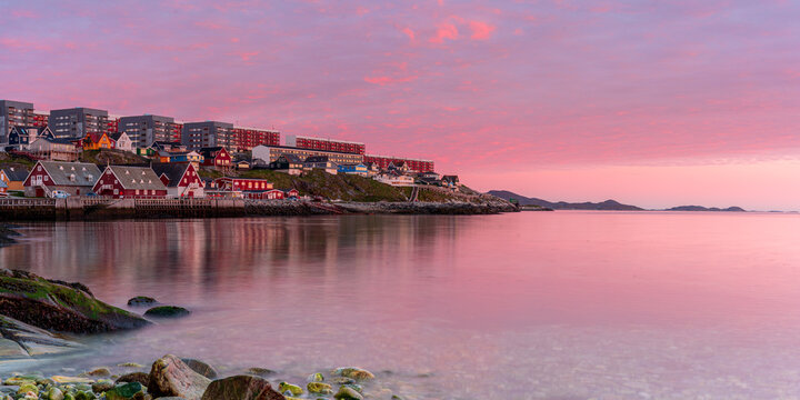 View of colorful houses along the rocky coastline under a vibrant pink sunset sky reflecting on the calm water of the harbor in Nuuk, Sermersooq, Greenland.