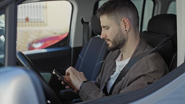 Man texting on smartphone while seated belted in parked car on street, looking down and tapping touchscreen with both hands, wearing jacket and white tshirt; focus.
