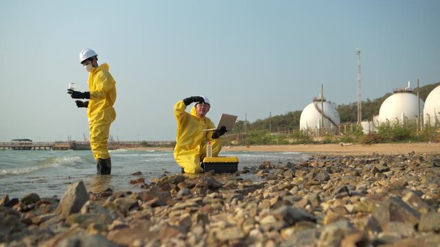 Two scientists in yellow suits are collecting water samples from the shore. One collects the water in a test tube, while the other analyzes and records preliminary data on a laptop before taking the w