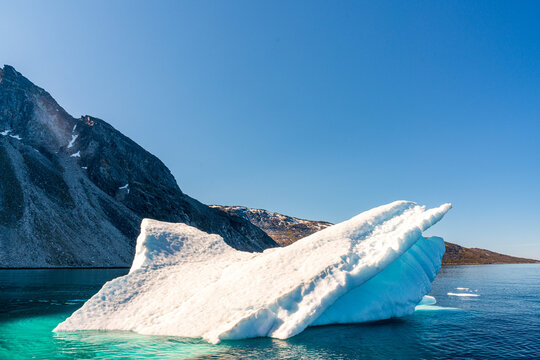 View of a large white iceberg floating in turquoise water with dark rocky mountains under a clear blue sky in Nuuk, Sermersooq Municipality, Greenland.
