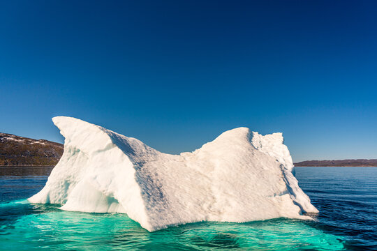 View of a massive white iceberg floating in the turquoise waters of a fjord with rocky hills under a clear blue sky Nuuk, Sermersooq Municipality, Greenland.