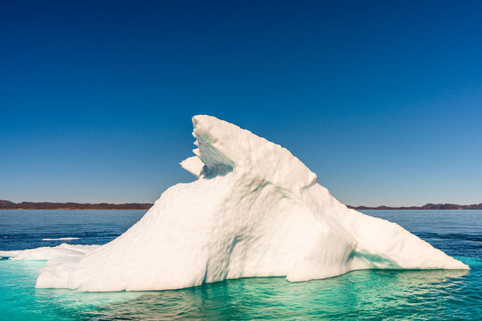 View of a massive white iceberg with jagged peaks floating in the deep blue sea under a clear sky with distant hills on the horizon Nuuk, Sermersooq Municipality, Greenland.