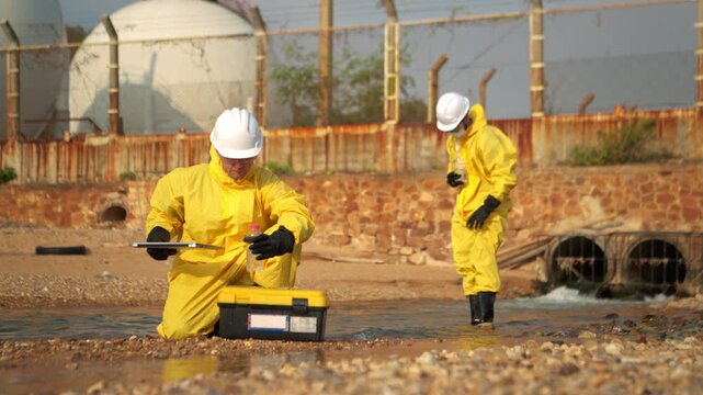 Two scientists in yellow suits are collecting water samples from the shore. One collects the water in a test tube, while the other analyzes and records preliminary data on a laptop before taking the w