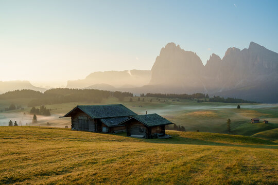 View of traditional wooden alpine huts on the rolling green meadows of Alpe di Siusi with the majestic Sassolungo mountain peaks under a soft morning mist in Trentino-South Tyrol, Italy.