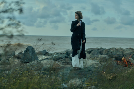 Outdoor fashion seascape on a windy rocky shore, coat and dress fluttering, capturing a moody windblown scene along the coast with dramatic silhouette, calm ocean and cloudy sky.