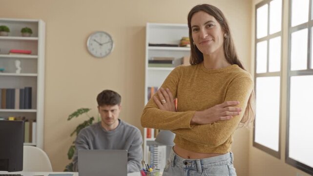 Woman standing at desk with arms crossed and smiling, laptop and bookshelf visible in office with clock and window; confidence collaboration.