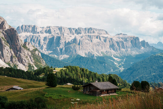 View of traditional wooden alpine huts nestled in green meadows with the jagged peaks of the Dolomites under a cloudy sky in Trentino-South Tyrol, Italy.