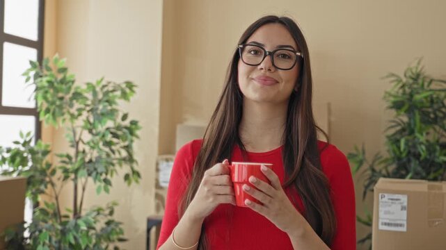 Woman holding red coffee mug and standing among delivery parcels and moving boxes in building; small business contentment.