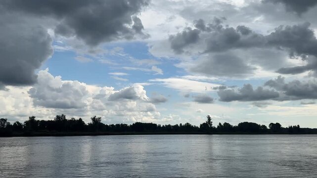 Dramatic summer sky over Siberian river with dark storm clouds and blue patches reflecting on calm water, lush tree silhouettes on far bank.