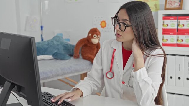 Young woman doctor wearing stethoscope types at computer while touching neck and glancing at screen in medical building; focused care.