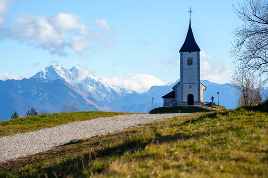 View of the Church of St. Primus and Felician in Jamnik on a grassy hill with snow-capped mountains in the background Slovenia.