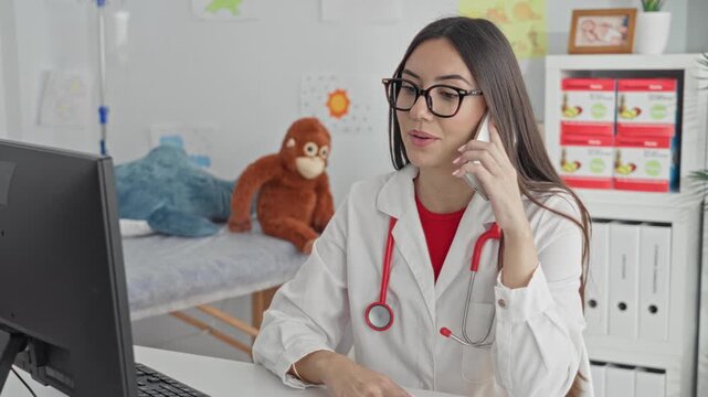 Young woman doctor holds phone to ear while gesturing with hand and using computer, red stethoscope visible in pediatric clinic; telehealth consultation compassion.