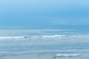 Fototapeta premium Calm ocean waves roll toward the shore beneath a pale blue sky, forming a tranquil coastal seascape of soft surf, gentle ripples and sea foam that conveys peace, relaxation and quiet reflection.
