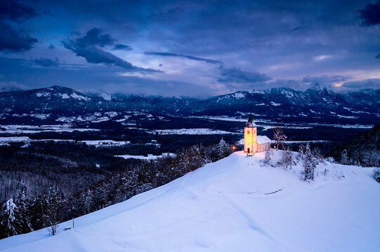 View of the illuminated Church of St. Primus and Felician on a snow-covered hilltop with the Julian Alps in the background at dusk in Slovenia.