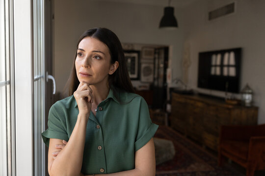Sad adult 30s woman standing alone by window in living room, looks outside with worried, reflective expression, thinking about life challenges, personal problems, loneliness or making decision at home