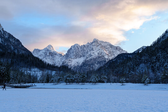 View of snow-capped mountain peaks and frozen Lake Jasna surrounded by evergreen trees under a sunset sky Slovenia.