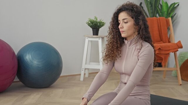Young woman reaching toes in a seated forward fold on a yoga mat, socks and activewear visible beside exercise balls and a stool in a studio; mindful calm.